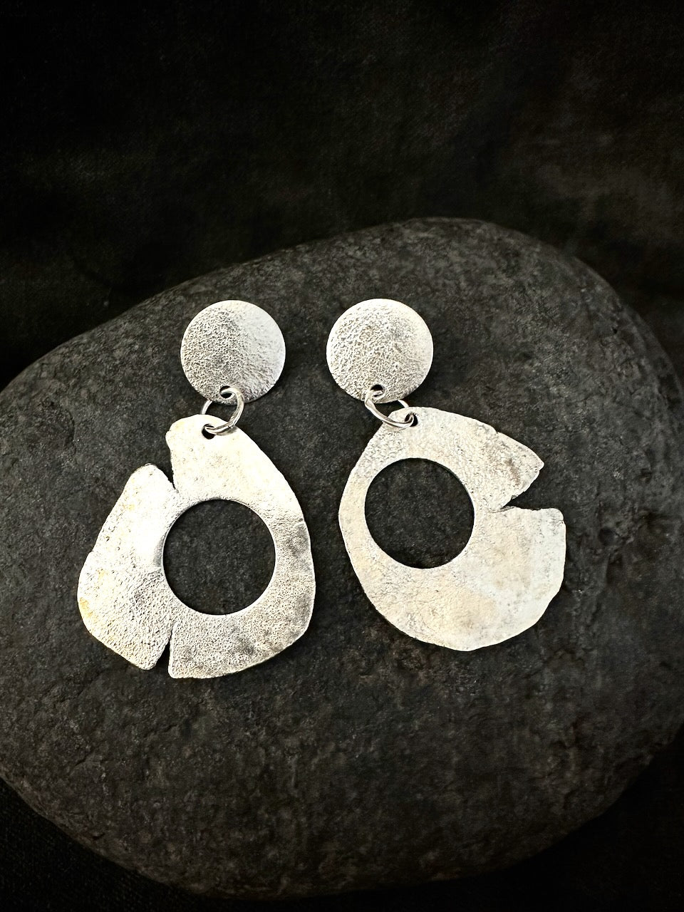Pair of sculptural silver drop earrings on dark stone. Recycled sterling silver, matte textured disc and large organic open hoop, handmade in France.