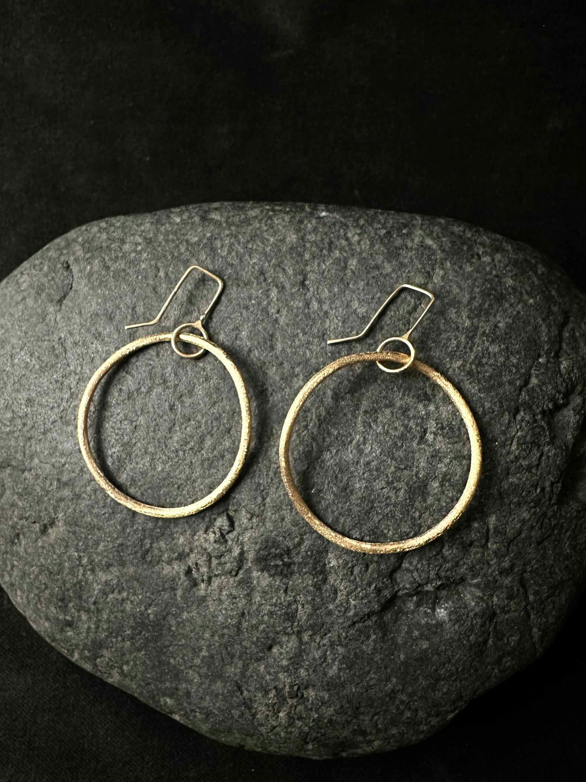 A pair of textured gold-tone hoop earrings with fishhook backings, displayed on a dark gray stone against a black background.