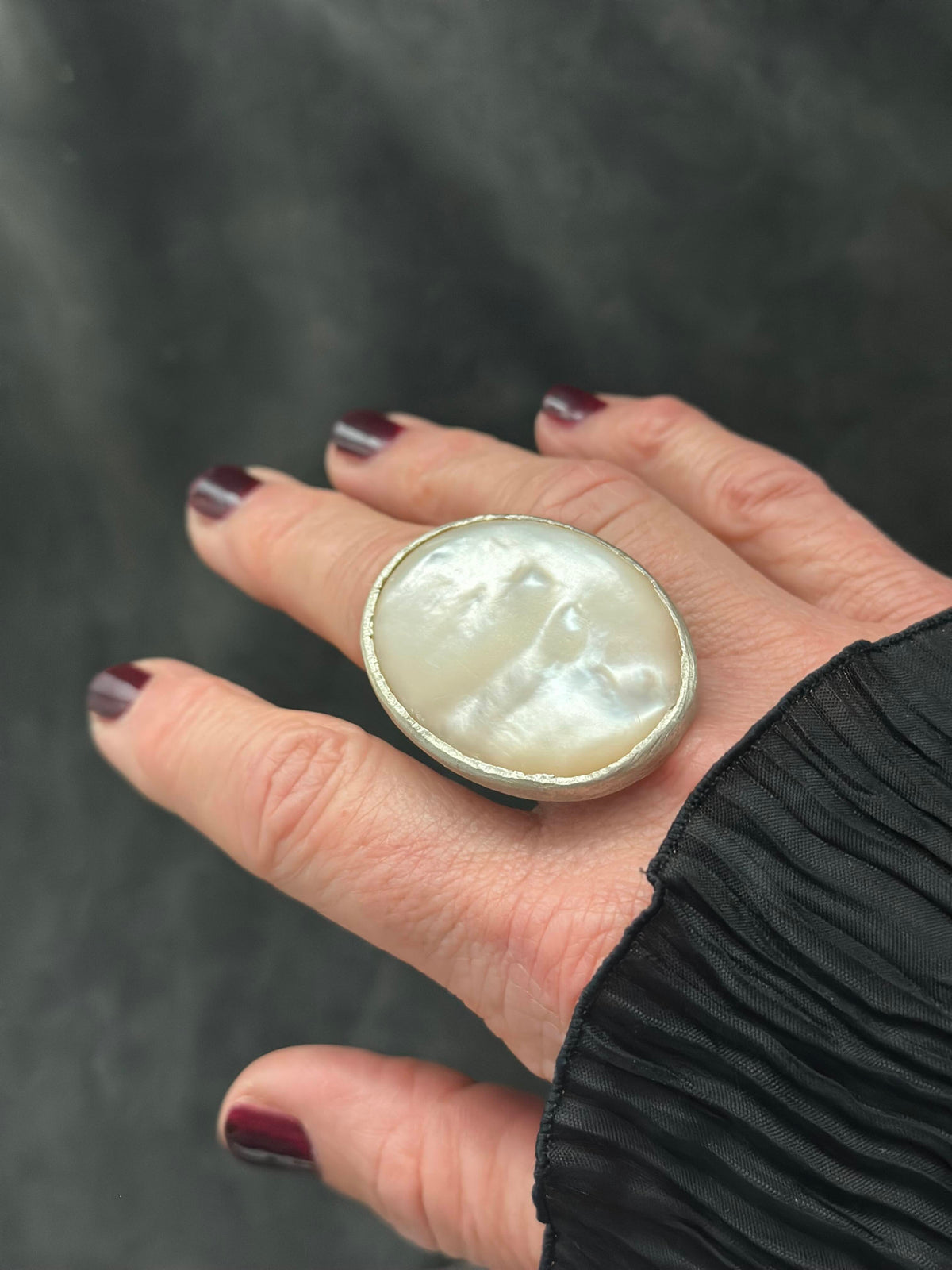 A close-up of a hand wearing a large oval ring with a milky white mother-of-pearl style face and a silver-toned bezel, against a dark gray textured background.