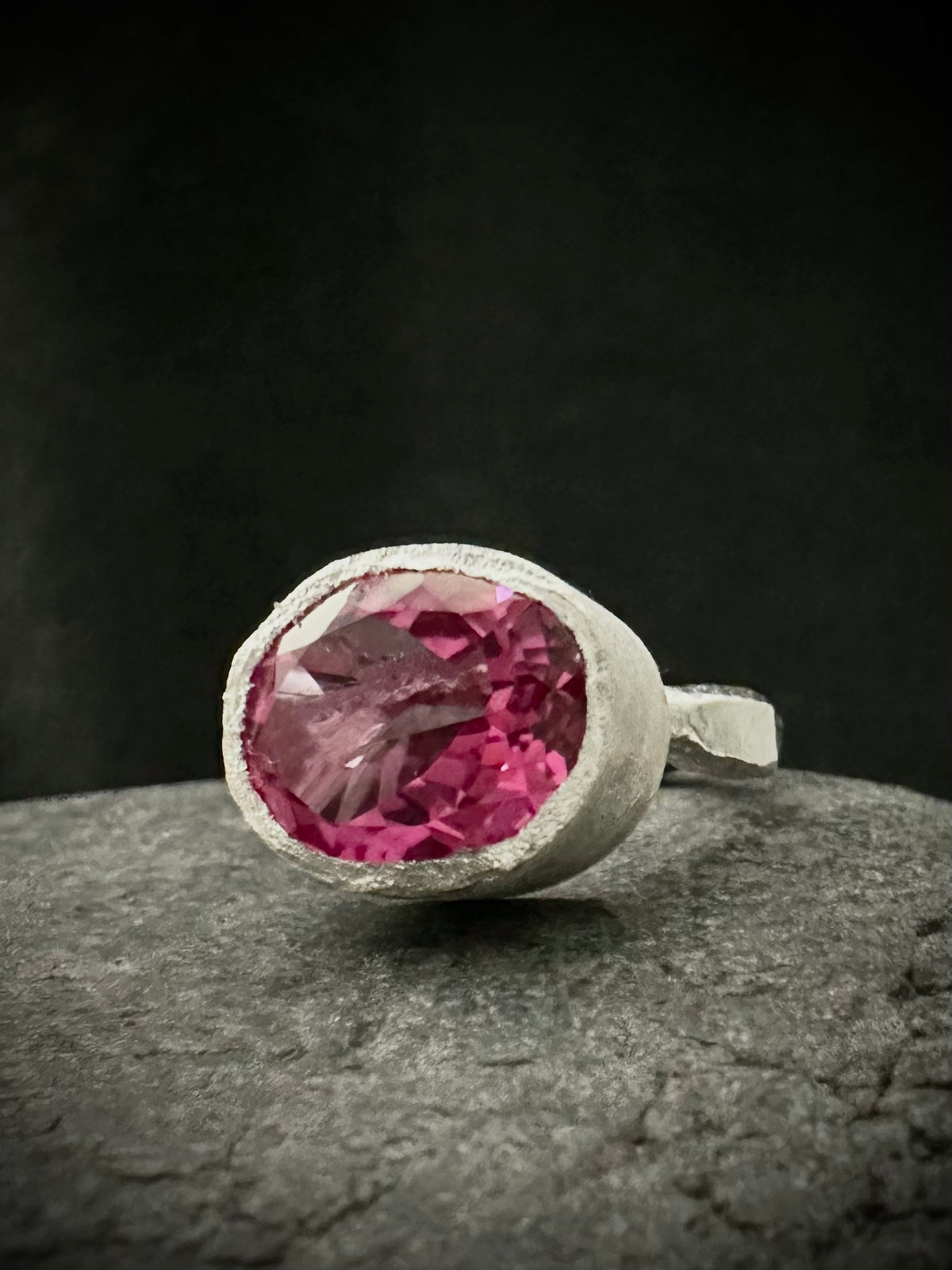 A silver ring featuring a large, oval-cut pink topaz gemstone in a textured bezel setting, resting on a dark gray stone surface against a dark background.