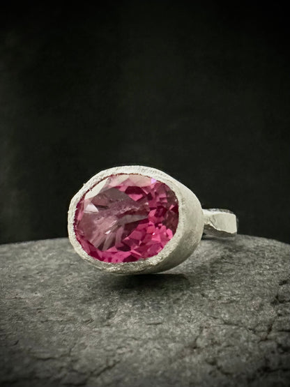 A silver ring featuring a large, oval-cut pink topaz gemstone in a textured bezel setting, resting on a dark gray stone surface against a dark background.
