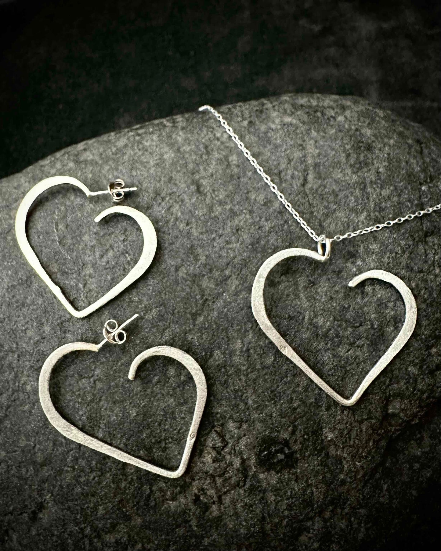 Sterling silver open heart jewellery set with handmade heart stud earrings and matching heart pendant necklace, photographed on dark stone background