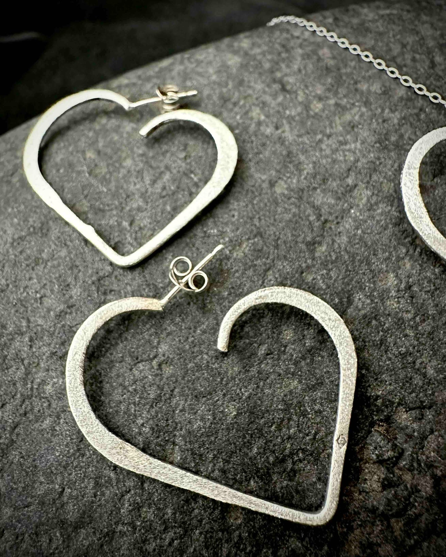 Handmade sterling silver heart stud earrings with hammered textured finish, minimalist open heart design on dark stone surface