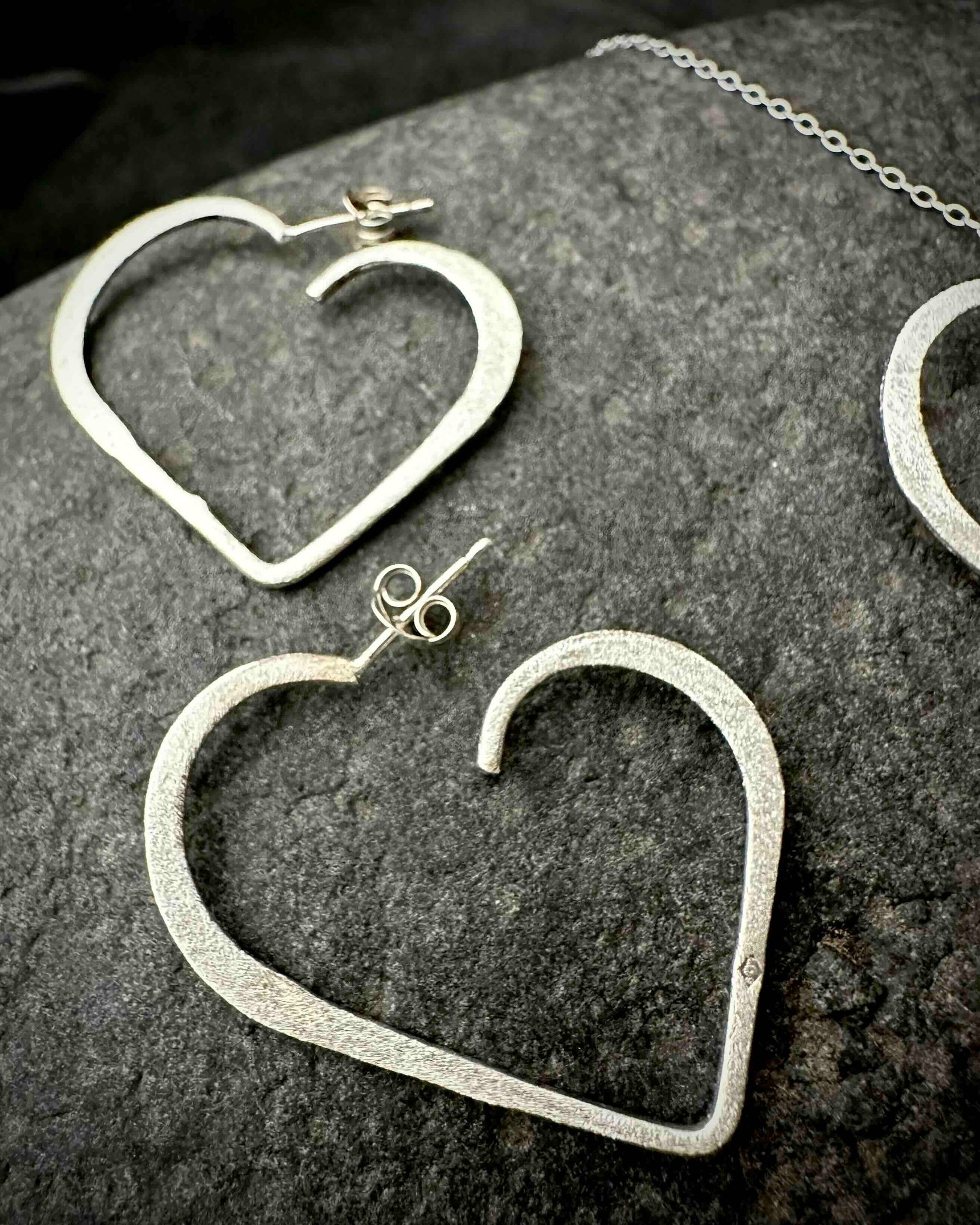 Handmade sterling silver heart stud earrings with hammered textured finish, minimalist open heart design on dark stone surface