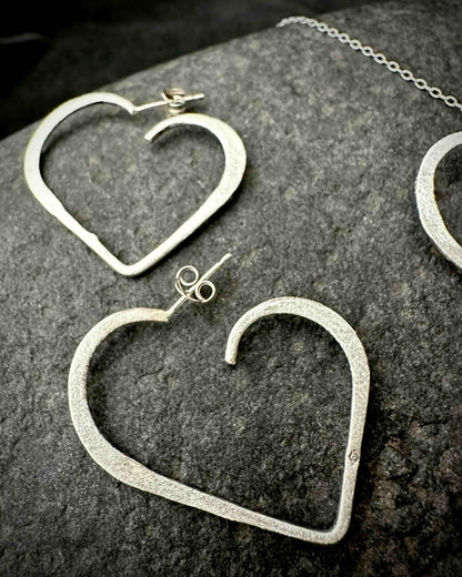 Handmade sterling silver heart stud earrings with hammered textured finish, minimalist open heart design on dark stone surface