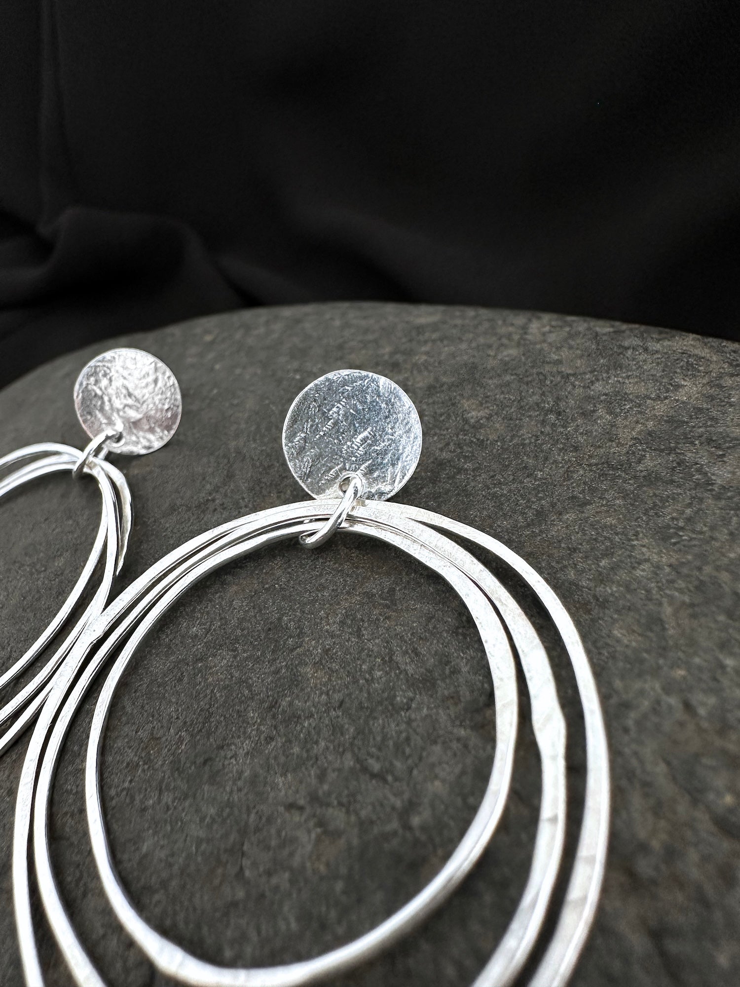 A pair of silver-toned dangle earrings with textured circular studs and layered thin hoop drops, displayed on a dark textured surface against a black fabric background.