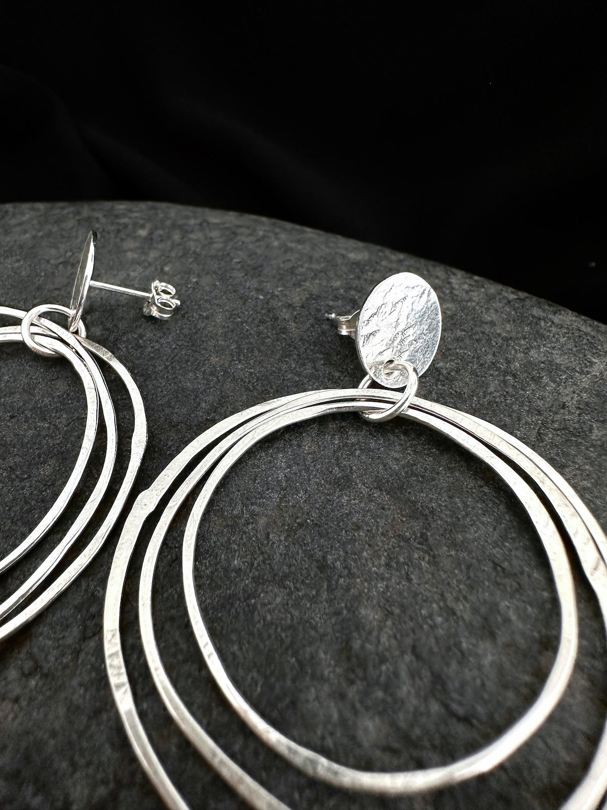 Sterling silver multi-ring hoop earrings with a textured oval disc charm, displayed on a dark textured stone surface.