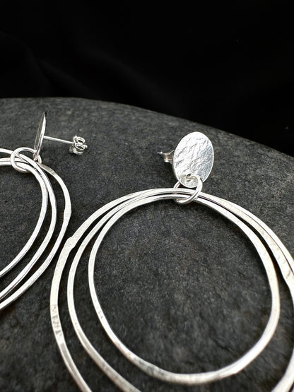 Sterling silver multi-ring hoop earrings with a textured oval disc charm, displayed on a dark textured stone surface.