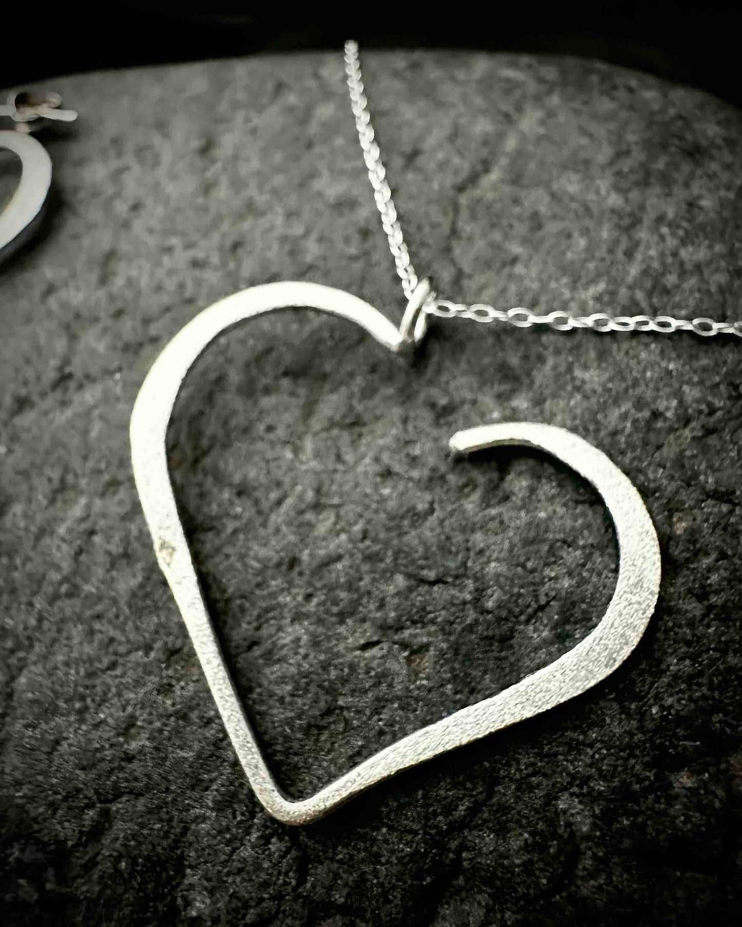 Handmade sterling silver heart stud earrings with hammered textured finish, minimalist open heart design on dark stone surface