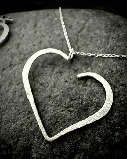 Handmade sterling silver heart stud earrings with hammered textured finish, minimalist open heart design on dark stone surface