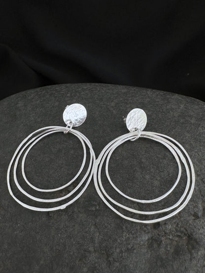 A pair of silver-toned triple-ring hoop earrings with textured circular studs, displayed on a dark gray stone against a black fabric background.