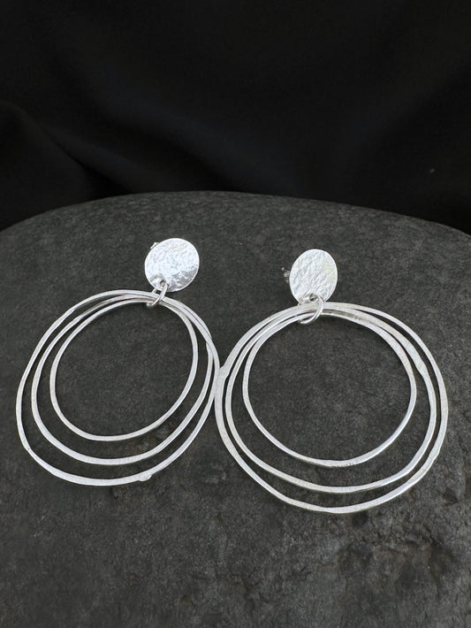 A pair of silver-toned triple-ring hoop earrings with textured circular studs, displayed on a dark gray stone against a black fabric background.